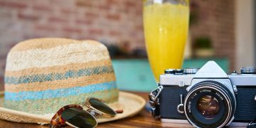 A straw hat, sunglasses, vintage camera, and a glass of orange juice on a wooden table.