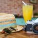 A straw hat, sunglasses, vintage camera, and a glass of orange juice on a wooden table.