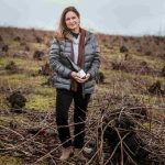 Woman standing in a field of bare, pruned vines on an overcast day, wearing a gray puffer jacket and brown scarf, holding a bottle.